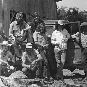 Group of long haired men and woman posing with truck and barn in the background