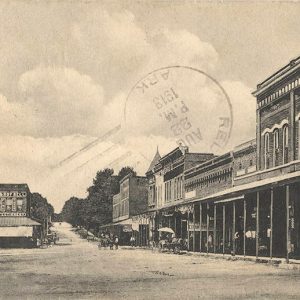 People and horse drawn carriages on dirt road between multistory brick buildings with covered entrances