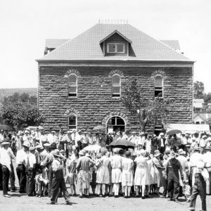 Two-story brick building with arched windows surrounded by a crowd of people, some with umbrellas