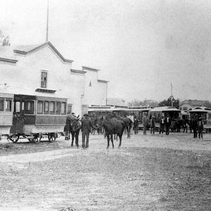 Men with horses and street cars outside a large white building