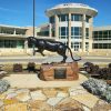 Panther statue on rock pedestal with plaque on platform in flower bed and multistory building with round entrance in the background