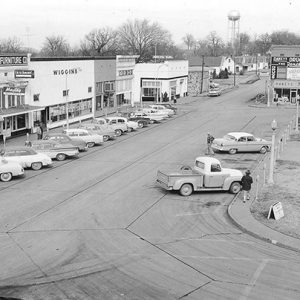 Town square with storefronts parked cars and water tower in distance