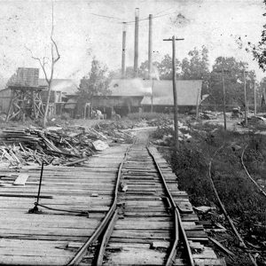 Factory buildings with smokestacks and pile of lumber next to railroad tracks in the foreground