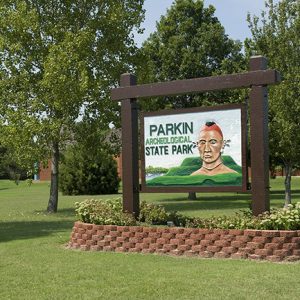 Wooden sign with Native American man painted on it and brick base with flowers and trees behind it