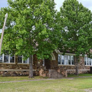 Single-story stone building with arched doorway and trees in front yard