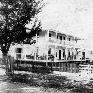 Two-story building with trees and outbuildings behind a fence