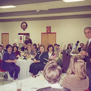 White man in suit and tie speaking at lectern to white men and women seated at tables