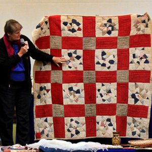 White woman talking into a microphone about a red and white quilt on stage