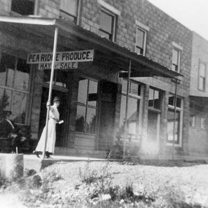 White woman in dress on sidewalk in front of two-story produce store