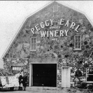 White man and white woman standing before large stone building "Peggy Earl Winery"