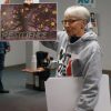 Older white woman with glasses in hooded sweater holding up a painting with the word "Resilience" on it