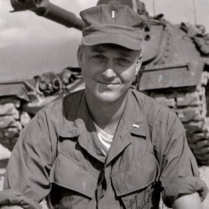 White man in military uniform with tank behind him