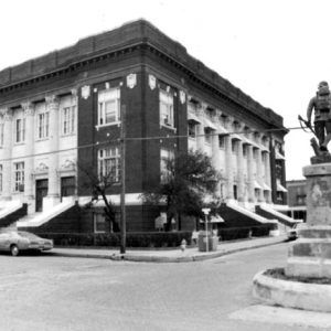 Multistory brick building with two sided greek facade and bronze soldier monument in street intersection