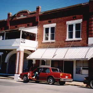 Two-story brick building with arched entrance way and covered balcony with red car in parking lot