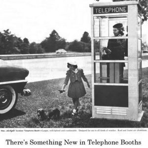 White woman using a telephone booth while watching her daughter walk the dog by their parked car on highway