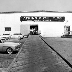 Group of white men and forklift exiting "Atkins Pickle Company" packing warehouse on parking lot with sign and cars