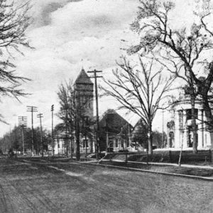 Multistory house with round covered porch and multistory building with tower on street with bare trees