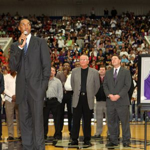 Tall African-American man in suit talking on microphone on basketball court with group standing on court behind him and crowd of spectators in background