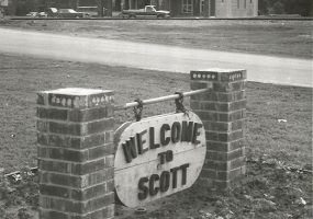 Multistory brick building with "Welcome to Scott" sign in the foreground