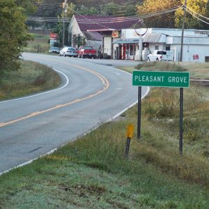 Single-story storefronts on two-lane street with "Pleasant Grove" sign in the foreground