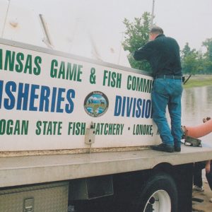 Two white men working with truck situated at the edge of a pond