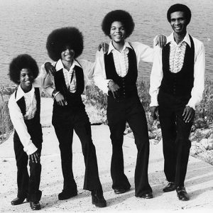 African-American man and three sons with afros and matching outfits of black pants and vests with white ruffled shirts standing together with body of water behind them