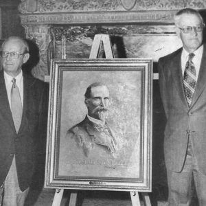 Pair of old white men with glasses in suit and tie standing at fireplace with framed painting of white man with beard in suit on easel in between them