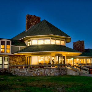 Multistory building lit up from the inside with brick chimneys stone walls and hexagonal covered porch on grass with walking path