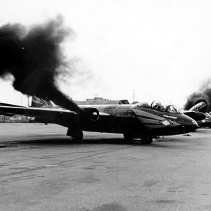 row of airplanes with first plane having dark smoke coming from its engines and more smoke coming from one of the other planes