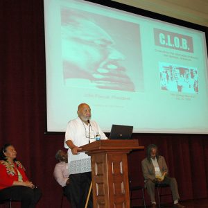 African-American man speaking at lectern with African-American woman and men sitting behind him on stage