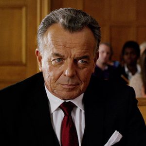 Older white man in suit sitting in court room with crowd sitting behind him