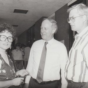 Pair of older white men speaking to white woman with glasses and vest in hallway