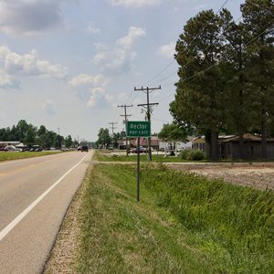 Two-lane road with "Rector" sign and single-story buildings in the background