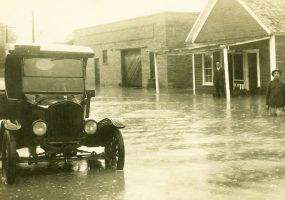 Black car parked on flooded street with two people standing in the waters in the distance