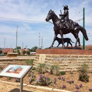 Bronze statue of man with hat and gun on a horse with dog on base in town with interpretation panel