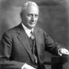 White man with white hair in three-piece suit posing in desk chair