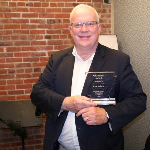 White man with glasses in suit standing holding up Arkansas shaped award with brick wall behind him