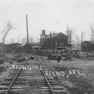 Railroad tracks with industrial buildings and water tower signed "cotton gins Reyno Ark"