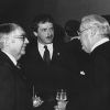 White man with mustache in suit and tie standing in between two older white men in suits with glasses talking