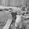 White child standing with logs and house behind him