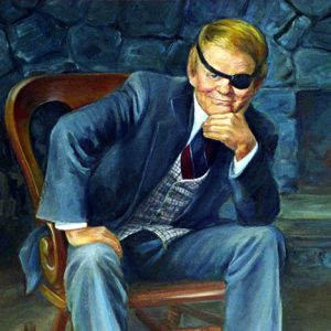 White man with eye patch in a suit sitting in a chair with fireplace