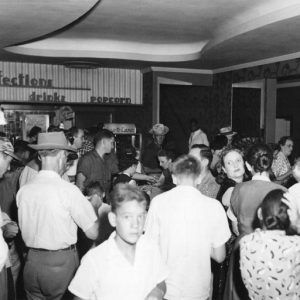 Crowd of white people in casual dress in theater lobby with wall sign "confections candy drinks popcorn"