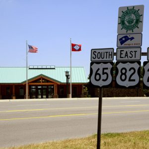 Road signs next to multilane street with single-story cabin building with green roof and flagpoles in the background