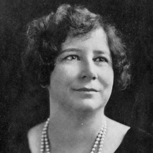 White woman with curly hair smiling in dress and two strings of pearls