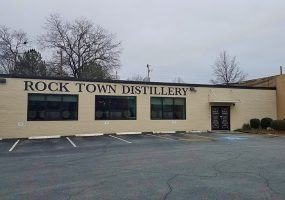 Single-story brick storefront with "Rock Town Distillery" painted above three large windows and parking lot