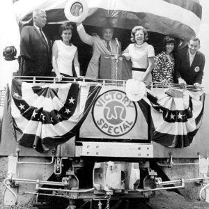 White men and women on the back of a train car labeled "W R Victory Special" with man in center standing in front of  a microphone and raising a cowboy hat in his right hand.