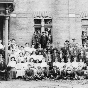 White male and female students and teachers pose for a group photo outside brick building with arched windows and covered entrance