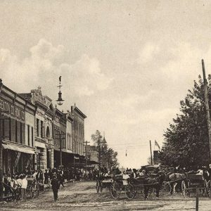 People and horse drawn carriages on crowded town street with multistory brick buildings on one side