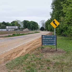 "Rohwer Japanese American Relocation Center" sign and yellow road sign on two-lane road with houses in the background