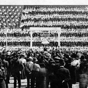 White man in suit speaking on stage with "human flag" in stands behind him and crowd around him